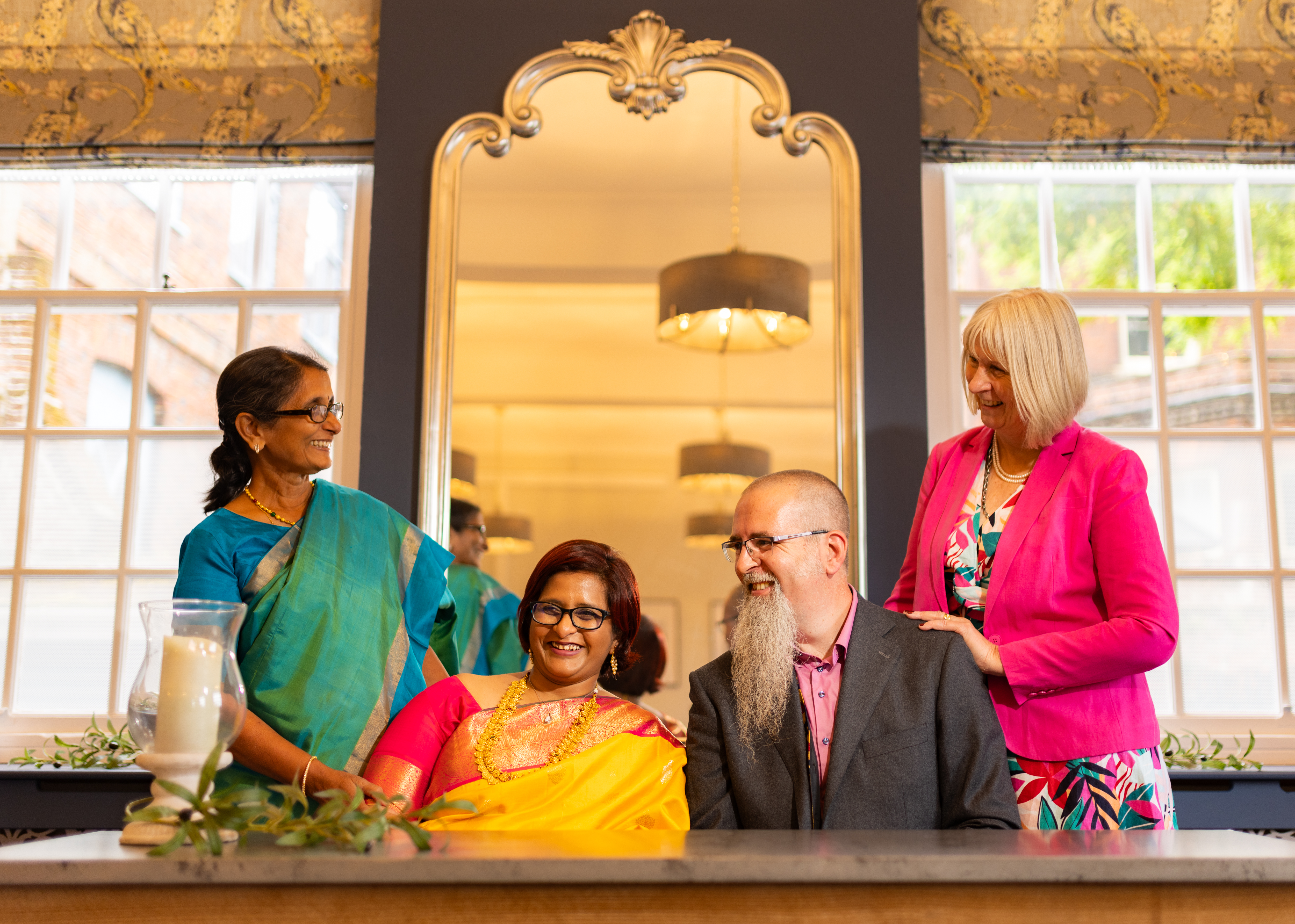 Bride in Indian dress sitting next to the groom with guests smiling either side of them.