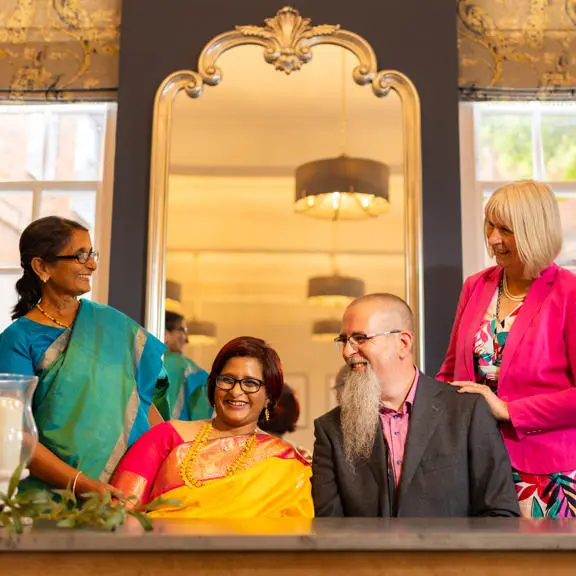 Bride in Indian dress sitting next to the groom with guests smiling either side of them.