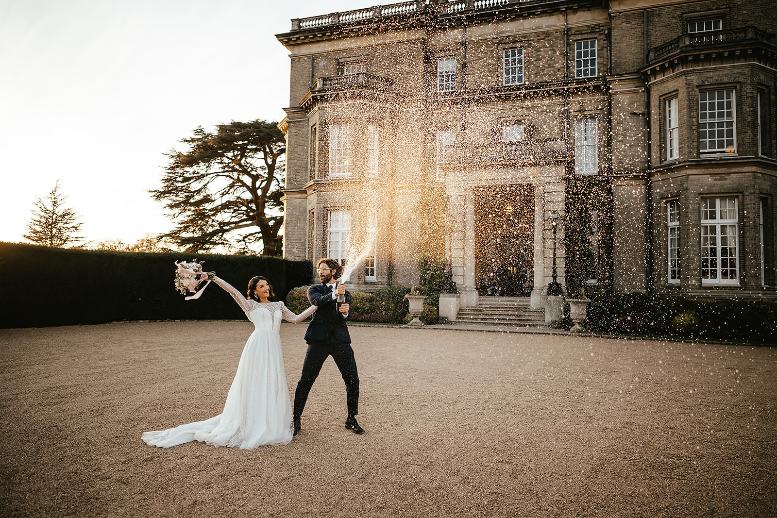 Bride and groom in front of grand mansion laughing and spraying champagne