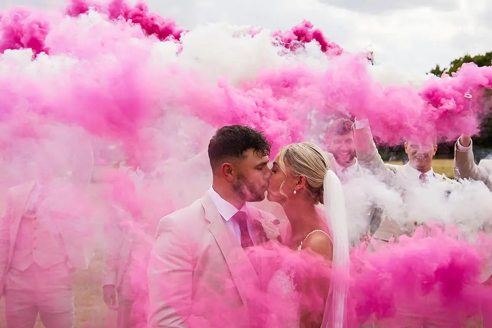 Bride and Groom kissing surrounded by friends amidst pink flares
