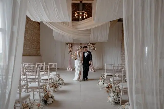 Bride and groom in beautifully decorated ceremony room with white chairs, white flowers and white drapery