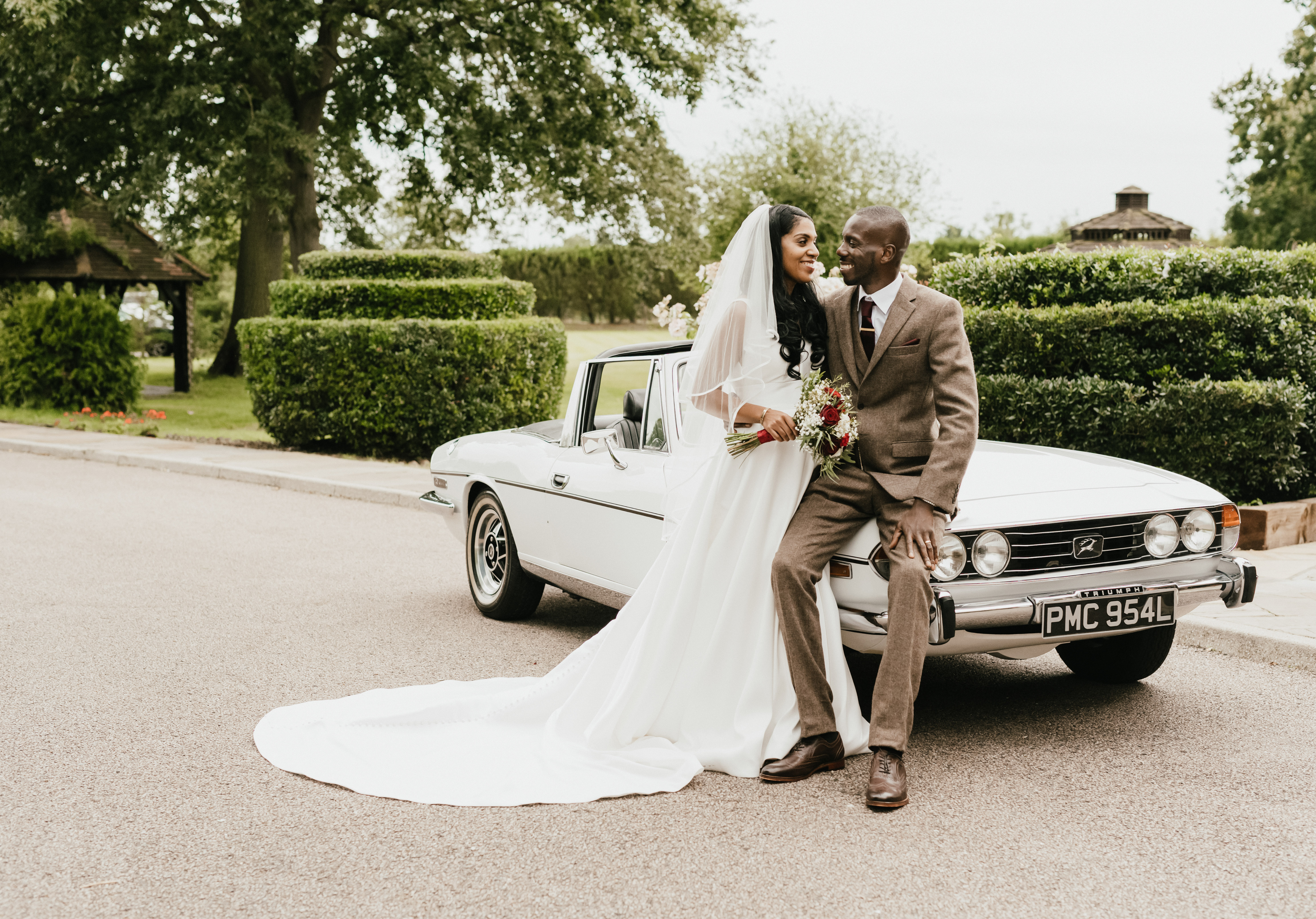 Bride and groom in parkland setting leaning against classic car
