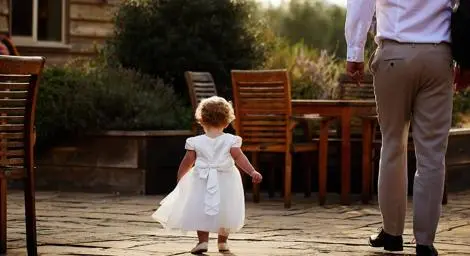 Little toddler girl in white dress at wedding walking with dad
