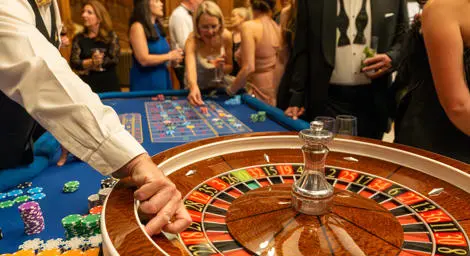 Roulette wheel with black tie guests in the background and showing part of croupiers arm releasing the ball