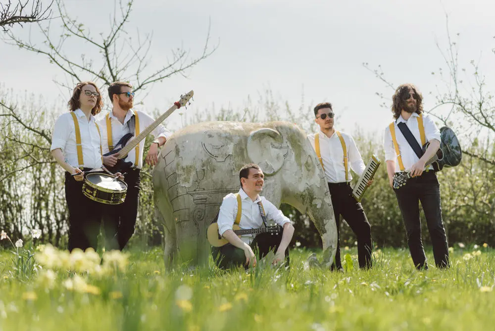 Five piece folk band dressed in white shirts and black trousers standing in meadow with a statue of an elephant