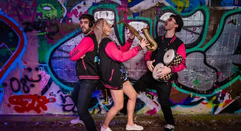 Three musicians in black pink jackets playing drums and saxophone in front of graffiti wall