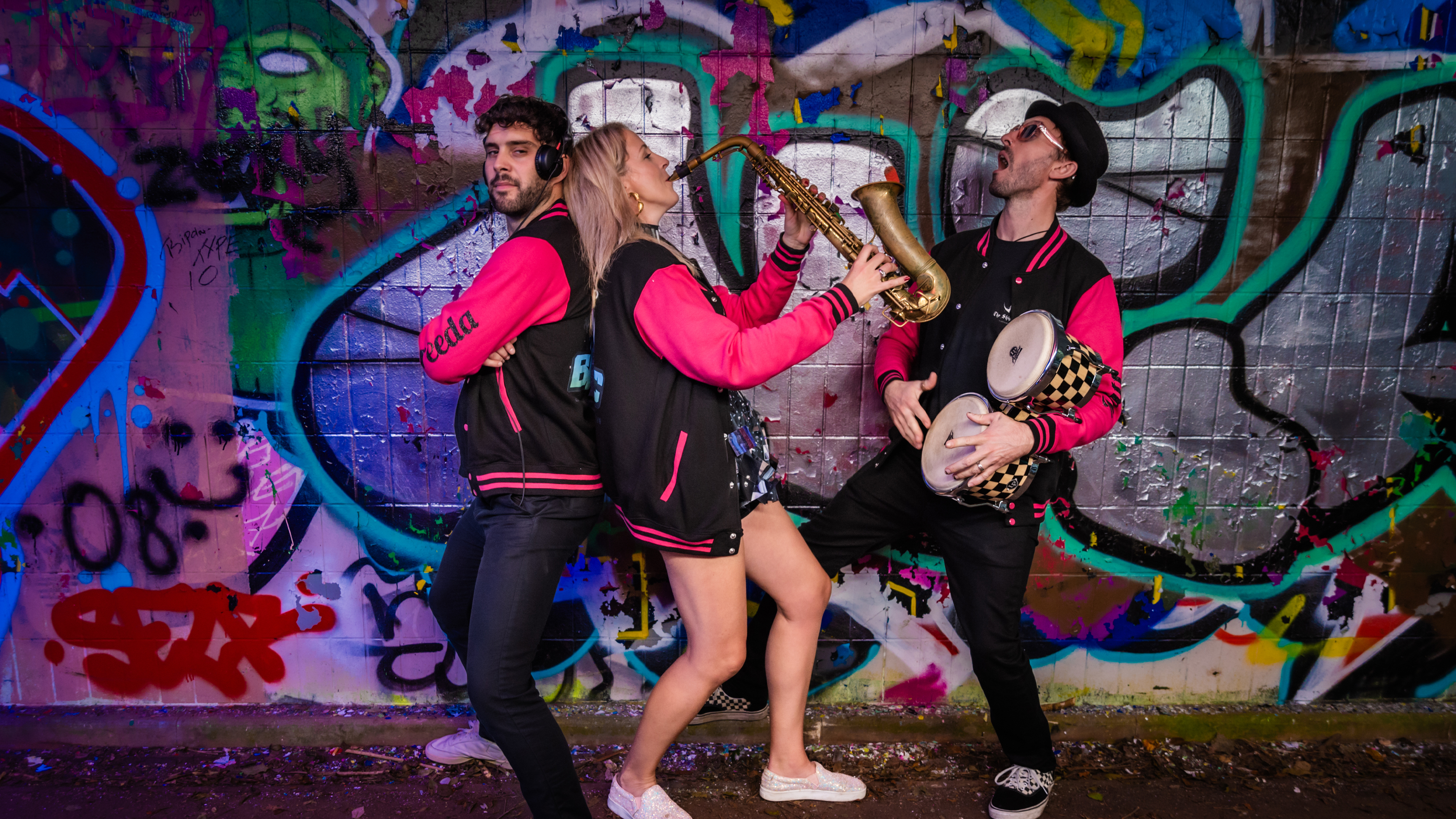 Three musicians in black pink jackets playing drums and saxophone in front of graffiti wall