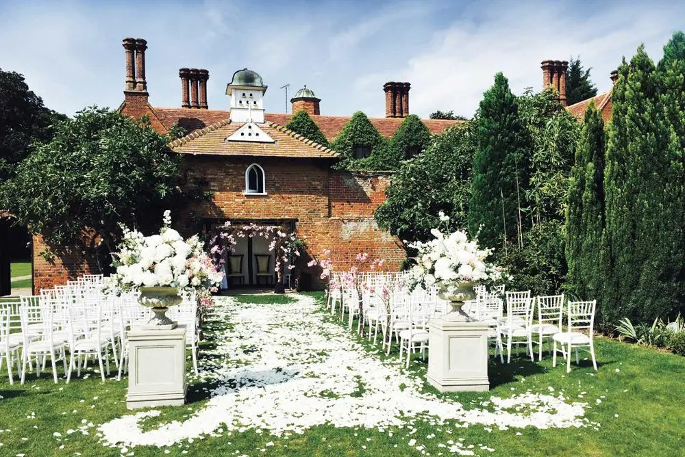 White chairs outside facing an arch with pink flowers. White roses either side of the aisle with petals scattered down.